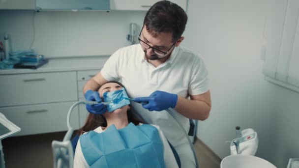 30 세의 남성 치과 의사가 안경을 끼고 치과 의자에 앉아 백인처럼 보이는 소녀의 치아를 치료하고 있습니다. 건강 한 미소 — 비디오