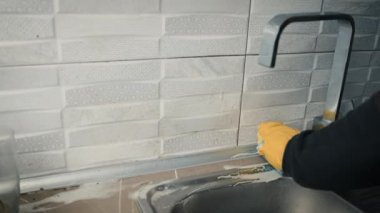 Sarı eldivenli bir elin yakın çekimi kirli seramik bir fayansı süngerle siliyor ve musluğun yanındaki mutfakta deterjan var. Temizlik ve temizlik