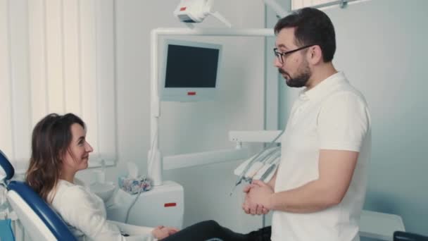 Ένας οδοντίατρος καυκάσιας εμφάνισης σε oulars επικοινωνεί με έναν ασθενή σε εκατό οδοντιατρική καρέκλα — Αρχείο Βίντεο