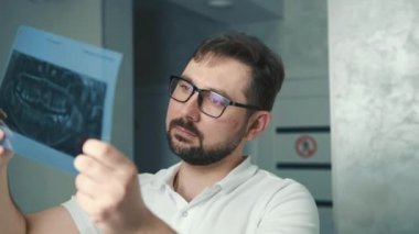 30 yaşında beyaz bir dişçi ışığın yanında duruyor ve hastaların röntgenlerine bakıyor.