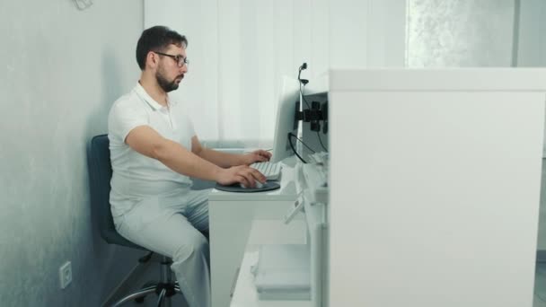 Biały dentysta siedzi przy komputerze w recepcji i nagrywa pacjenta. — Wideo stockowe