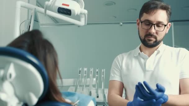 Kaukasischer Zahnarzt kommuniziert mit dem Patienten auf dem Zahnarztstuhl und zieht blaue Handschuhe an, bevor er einen Zahn füllt — Stockvideo