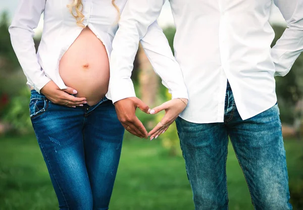 The concept of pregnancy, love, family and children. A pregnant woman in a white shirt with her husband makes a heart symbol with her hands.