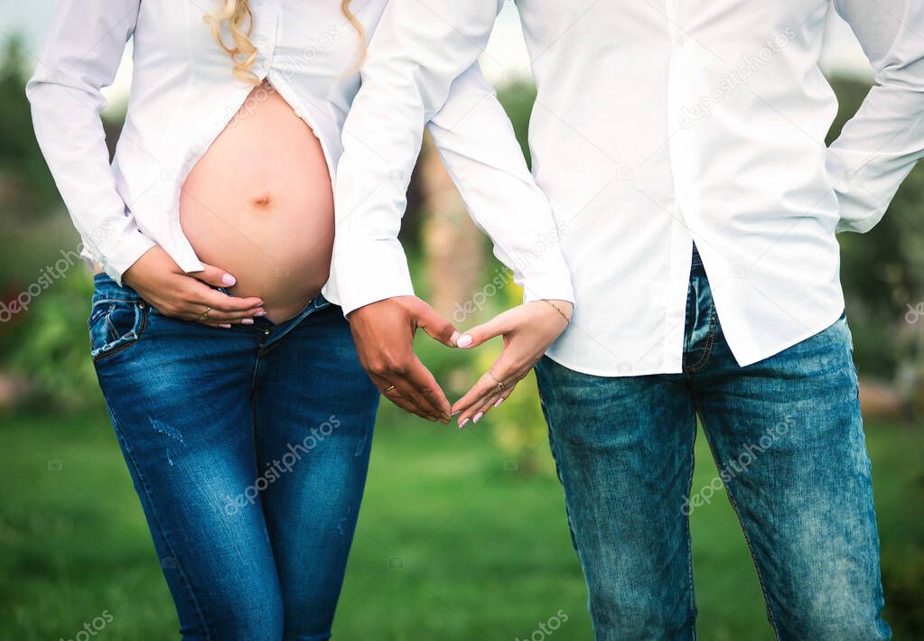 The concept of pregnancy, love, family and children. A pregnant woman in a white shirt with her husband makes a heart symbol with her hands.