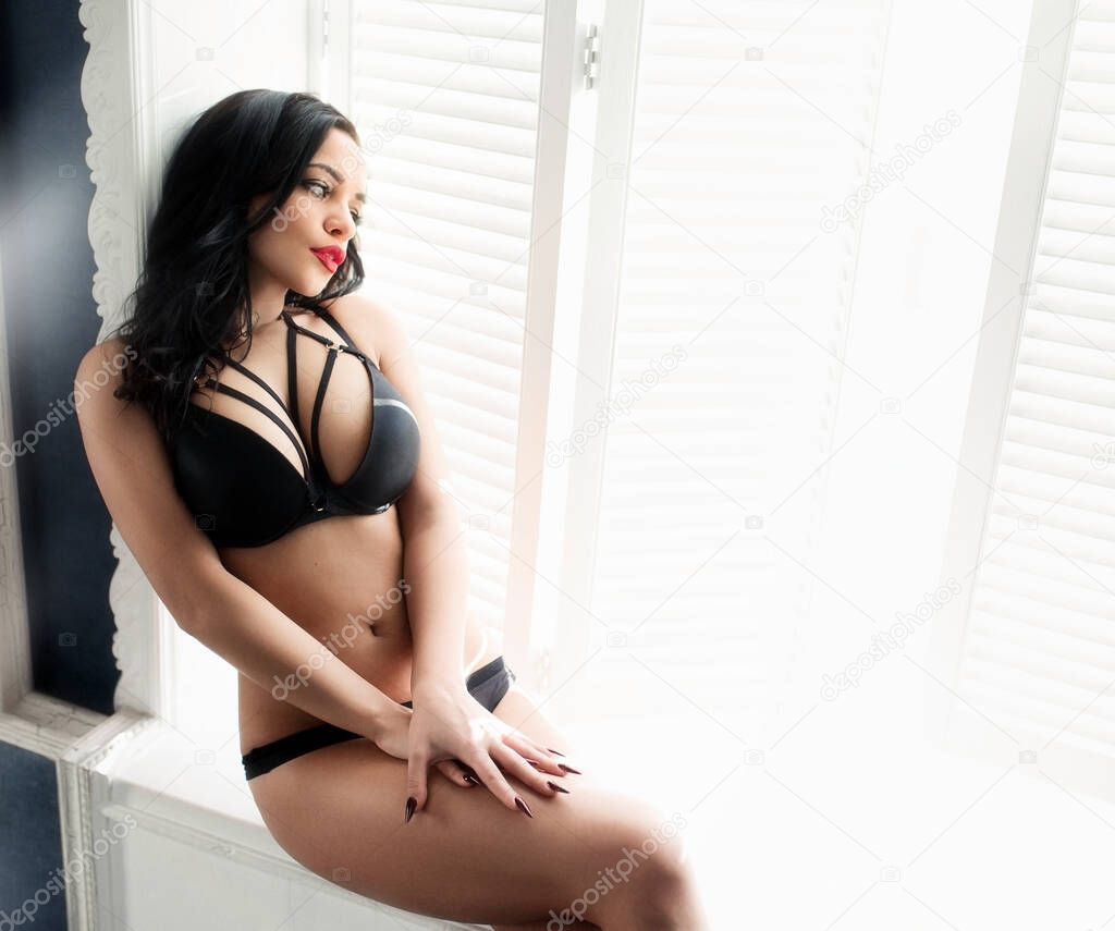 A young model in black lingerie with a tanned body poses sitting by the window. The concept of women beauty and sexy underwear.