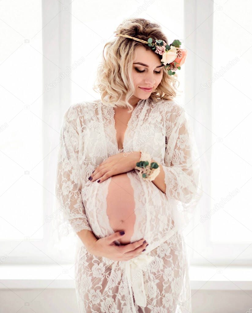 A beautiful pregnant woman with flowers stands at the window in the room.