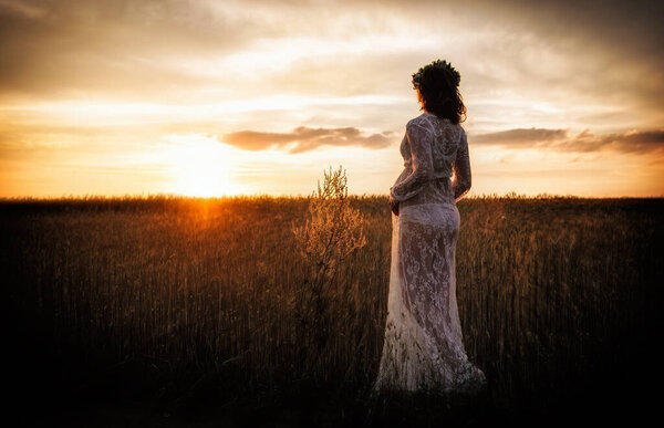 A pregnant woman in a delicate negligee in a field enjoys a beautiful sunset. Beauty, health and tender motherhood.