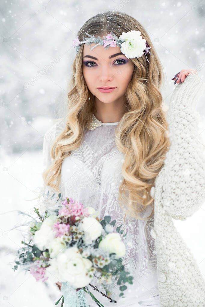 Attractive winter girl in a white outfit in a snow forest with beautiful flowers on her head and a bouquet. Soft focus and blur effect.
