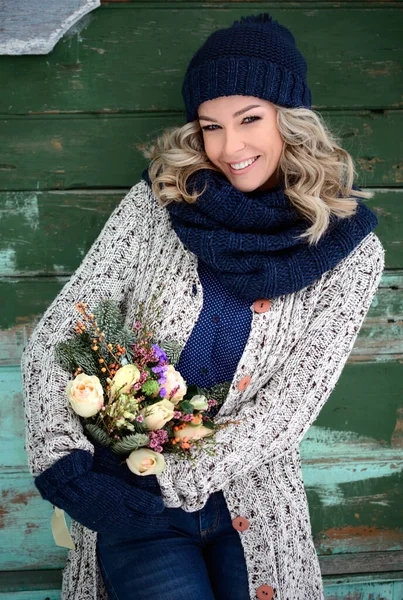 An attractive woman with a perfect smile stands against the wall and holds a bouquet of roses and fir branches. Stylish winter clothing