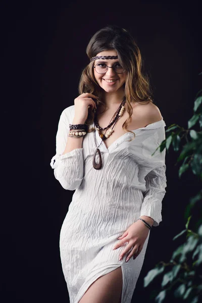 Beautiful fashionable smiling woman in a white dress with boho accessories. An attractive model with hippie style jewelry on a dark background