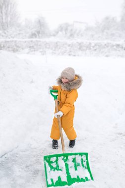 Turuncu tulumlu kız büyük kar küreğini temizliyor. Şiddetli kar yağışı sonrası kar temizleme. Çocuk kar yığınlarını temizlemekten mutlu.