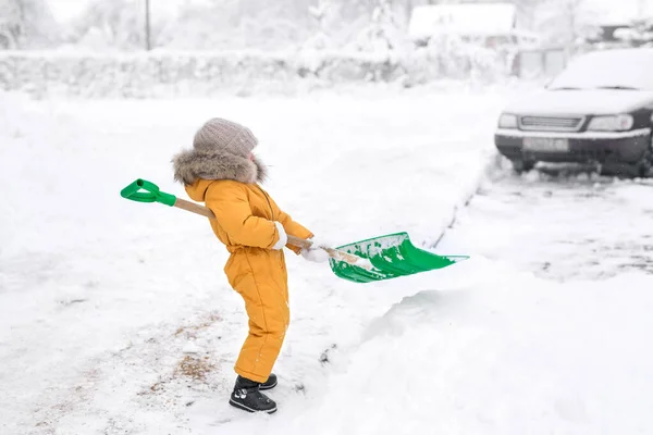 Girl in orange jumpsuit cleans snow big shovel. Snow removal after heavy snowfall. a child with difficulty lifts a shovel with snow from a snowdrift