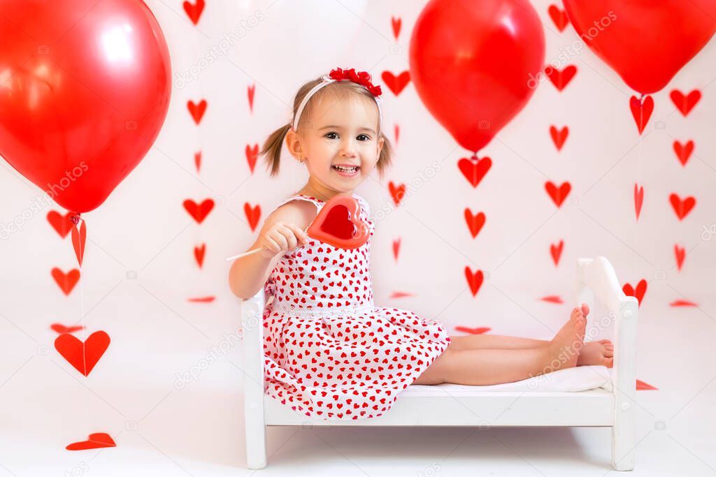 Baby holds red balls for Valentine's Day. A girl is eating a candy in the shape of a heart. A child in a white dress with hearts.