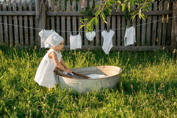The child merrily washes his clothes in the basin. Splashes of water and foam from washing clothes. A girl in a white dress hangs out wet underwear to dry. Washing in an old basin.