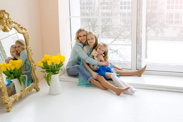 Mom hugs her daughters sitting at the window. A bouquet of yellow tulips in a vase stands by the window. Happy family. Flowers from daughters for mom on Mother\'s Day. child\'s day.