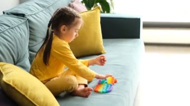 Küçük bir kız modern bir popit oyuncağıyla oynuyor. Etkileyici bir duyusal oyuncak. Renkli ve parlak bir oyuncak. Basit bir gamze. Çocuk sarı yastıklı bir kanepede oturuyor. Heyecan verici bir oyun
