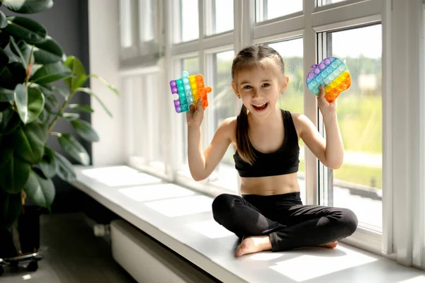 Kız büyük pencerenin yanındaki pencere pervazında oturuyor ve onu patlatma oyuncağıyla oynuyor. Ellerin iyi motor becerilerini geliştirmek için modern dokunmalı oyuncak popit. Basit bir gamze. Mutlu bebek