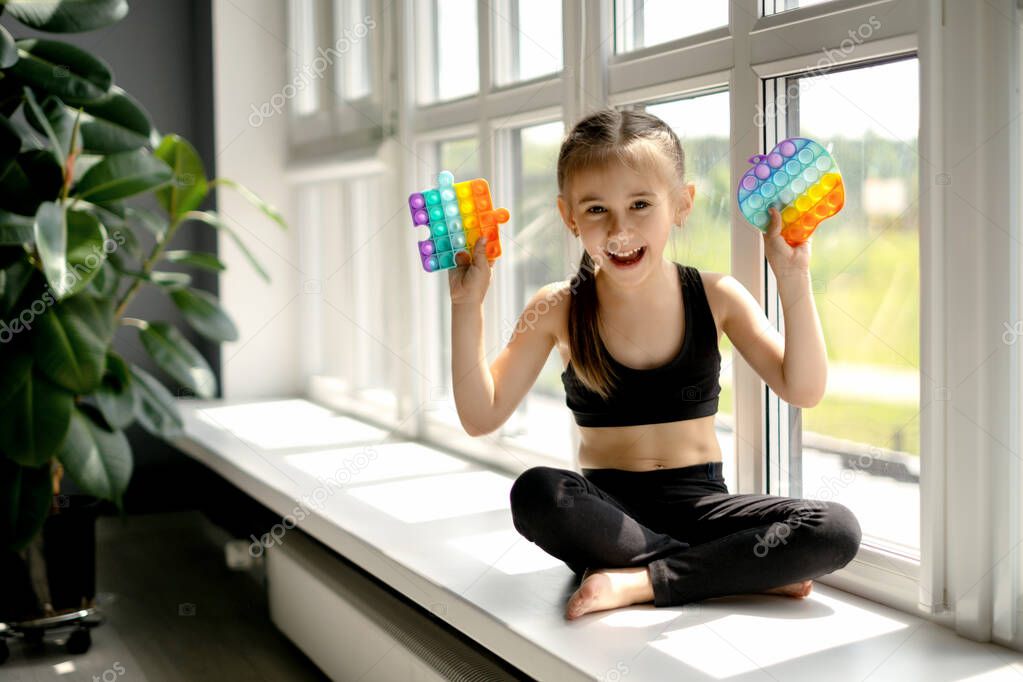 The girl is sitting on the windowsill by the big window and playing with pop it toy. Modern touch toy popit for the development of fine motor skills of the hands. simple dimple. Happy baby