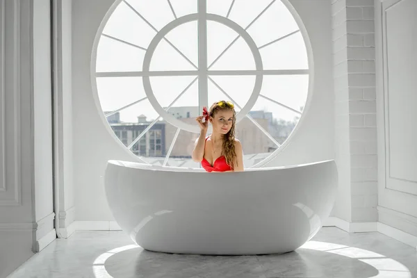 大きな丸い窓のある豪華な白いバスルームで花を持つ若いセクシーな女性 太陽の光が広々とした部屋を照らす 水着を着た赤いガーベラの女性 — ストック写真