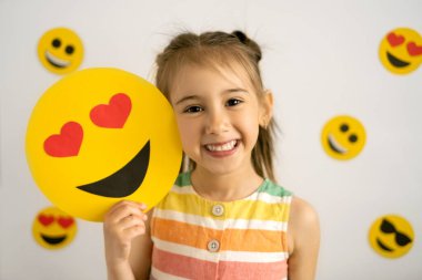 Dünya emoji günü. Antropomorfik gülen surat. Doğum günü partisi. Duygular. Küçük bir kız, tüm dişleriyle gülümsüyor, elinde bir karton aşk emojisi tutuyor..
