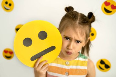 Dünya emoji günü. Antropomorfik gülümseme. Küçük bir kız, somurtkan üzgün, elinde karton üzgün bir emoji tutuyor. Değişik duygusal durumlara sahip emojilerle arka plan: üzgün, aşık, neşeli, mutlu