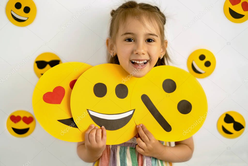 The girl holds cardboard smile faces with different emotions in her hands: a sad, smiling happy smile, a loving smile with hearts instead of eyes. A joyful little emotional girl. World emoji Day