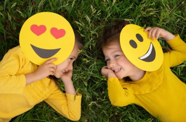 Two funny little girls are lying on the lawn covering part of their faces with emoticons and laughing merrily. World emoji Day. Anthropomorphic smile Face.