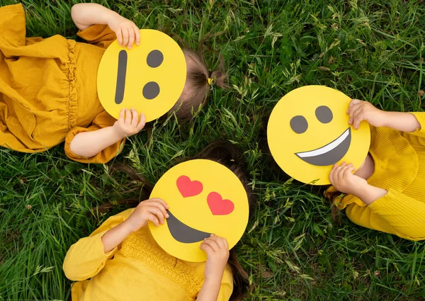Three children lying on the grass are holding cardboard emoticons with different emotions in their hands: a sad, smiling happy smile, a loving smile with hearts instead of eyes. World Emoji Day