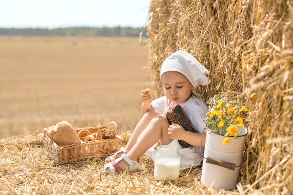 A girl in a white scarf is sitting in a field on a haystack and eating bread, washing down with fresh milk from a jar. breakfast in nature.healthy and full nutrition for children and adults.copy spase