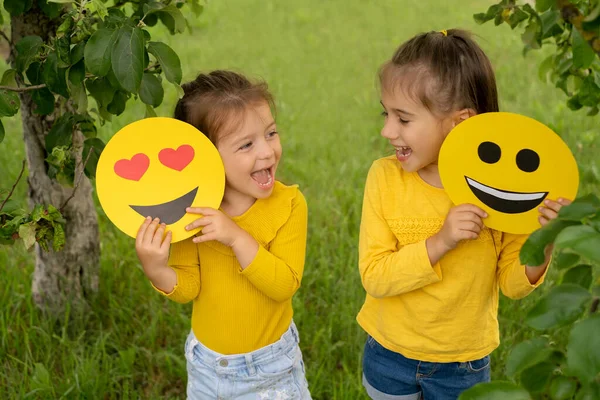 Two little funny girls are holding smile faces with different emotions in their hands. Children are having fun in the park. World emoji Day. Anthropomorphic smile.