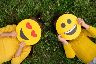 Two children hide behind the faces of cardboard funny happy emoticons lying on the lawn. Top view. World smile day