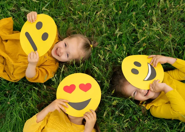 Three funny happy girls hide and look out from behind the faces of emoticons with different emotions : happy, sad and in love. Top view. Children are lying on the grass looking up. Emoji smile day