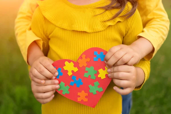 Two sisters are holding a red heart with colorful puzzles inside, made with their own hands as a sign of support for children with autism. World Autism Awareness Day. handmade