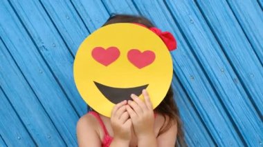 Sevimli küçük bir kız yüzünü karton bir gülen yüzle kapatıyor gözleri yerine kalpleri olan. Dünya emoji günü. Antropomorfik gülümseme. Duygular.