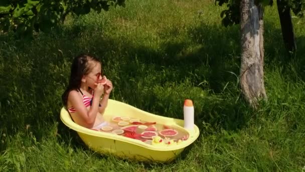 一个小女孩一边吃柚子 一边在户外洗澡时用柑橘类水果的碎片装满水 一个穿着泳衣 戴着红色太阳镜的孩子在水池里飞溅 — 图库视频影像
