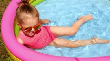 Güneş gözlüklü bir kız bahçedeki şişme evde eğleniyor. Sıcak bir yaz gününde bahçede çocukların dinlenmesi. Su sıçrıyor. Çocukların gülüşü. Küçük yuvarlak çocuk yüzme havuzu