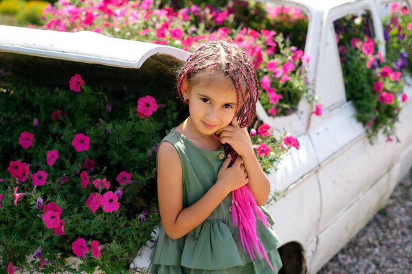 A girl in a dress with pink zizi pigtails from kanekalon stands at a decorative car with flowers growing inside. A kid at a flower bed with flowers. A modern child with a hippie hairstyle