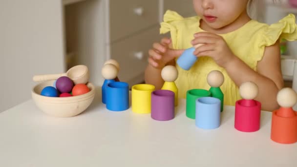 一个两岁的小女孩正坐在一张桌子旁 玩着一个形似男人的五颜六色木制圆筒 幼儿特写教育玩具分拣员 装有杯子和塑像 — 图库视频影像