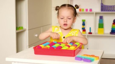 İki yaşındaki bir kız, oyun alanından renkli mozaik fayanslar topluyor. Çocuk odasındaki masada bir çocuk oyun oynuyor. Anaokulları için eğitici oyuncaklar. Bir çocuktan renk öğrenmek