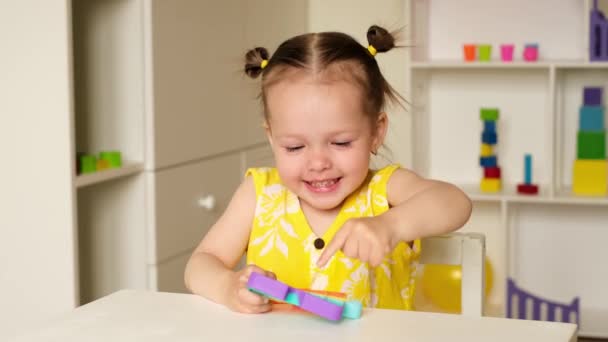 2歳の女の子がモダンなポピットおもちゃで子供部屋のテーブルで遊んでいます 開発のためのエキサイティングな感覚のおもちゃ 多色で明るいポップそれおもちゃ 子供のための便利なおもちゃ — ストック動画