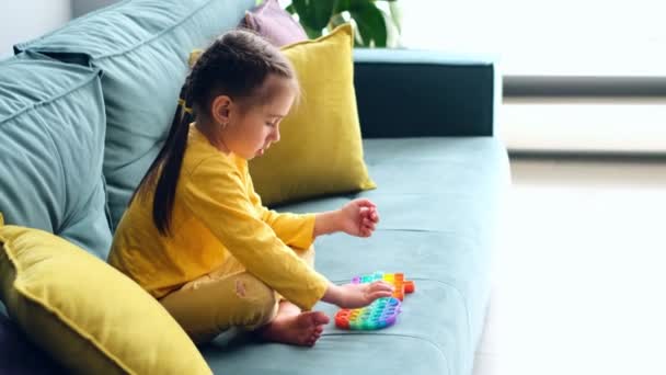 5歳の女の子がソファの上の部屋でおもちゃのおもちゃで遊んでいる 優れた運動能力の開発のための現代的な感覚玩具Ppt 子供たちのエキサイティングなゲーム 黄色の枕付きの緑のソファ — ストック動画