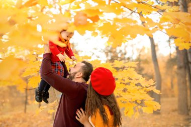 Baba, gülen kızını akçaağaç dallarının portakal yapraklarına fırlatır. Mutlu bir aile küçük bir kız büyütüyor. Kırmızı ve turuncu renkte giyin.