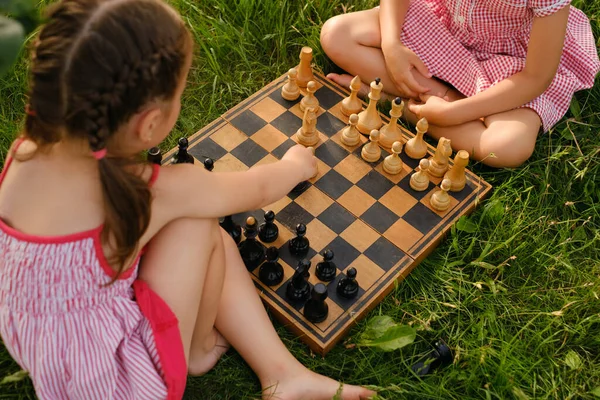 Little girls learn to play chess in the garden on an old chess board. Games for the development of thinking. The child makes a pawn move. Entertainment on vacation.