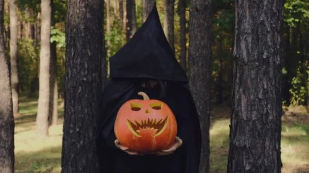 黒いレインコートを着たリーパーが木の幹の間の暗い森の中に立っています ハロウィーンのお祝いの前夜 トリックや治療 — ストック動画