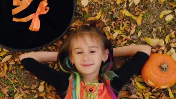 魔女の衣装を着た女の子が カボチャと帽子の隣にある秋の葉の地面に横たわっています トリックや治療 ジャック オランタン ハロウィーンの秋の季節割引と販売 — ストック動画