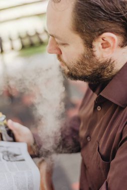 Kahverengi gömlekli bir adam gazete ve sigara okuyor.