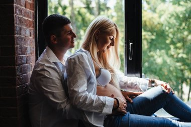 Mutlu adam büyük bir pencerede oturan hamile karısına sarılıyor. Yeni evlerinin pencere eşiğinde oturan mutlu genç bir aile. Sevgililer birlikte vakit geçirir. Dokuz ay.