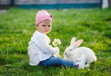 Küçük güzel kız bahçede bir tavşanla oynuyor. Güneşli bir günde yeşil çimenlerin üzerinde şirin bir bebek. Açık havada bir yaşındaki bir bebeğin portresi. Paskalya tatili konsepti. Paskalya tavşanı.