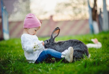 Küçük güzel kız bahçede bir tavşanla oynuyor. Güneşli bir günde yeşil çimenlerin üzerinde şirin bir bebek. Açık havada bir yaşındaki bir bebeğin portresi. Paskalya tatili konsepti. Paskalya tavşanı.