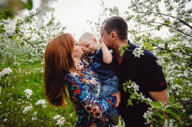 Mutlu ebeveynler ve bebek tatili. Bahar bahçesinde yürüyüş yapan mutlu bir aile, park. Anne, baba ve oğul portresi. Babam bir yaşındaki oğlunu kollarının arasına aldı. Aile tatili.